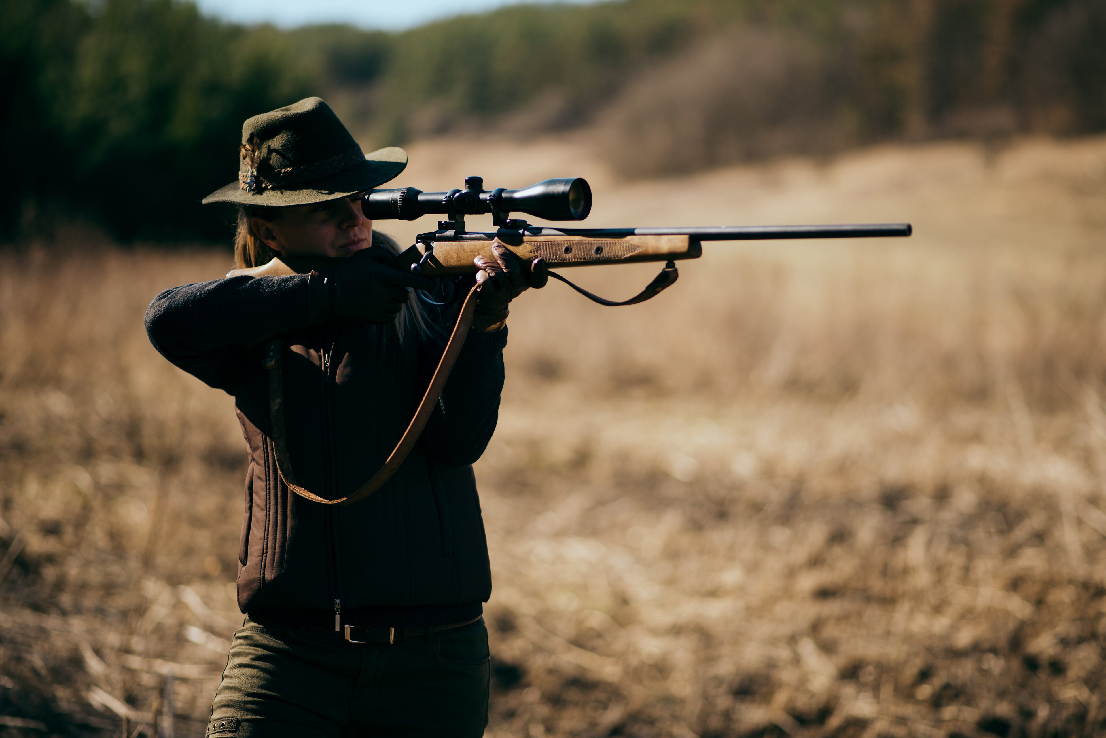 Woman wearing a hat aiming a scoped long rifle in an open field with green trees in the background–outdoor shooting scene.